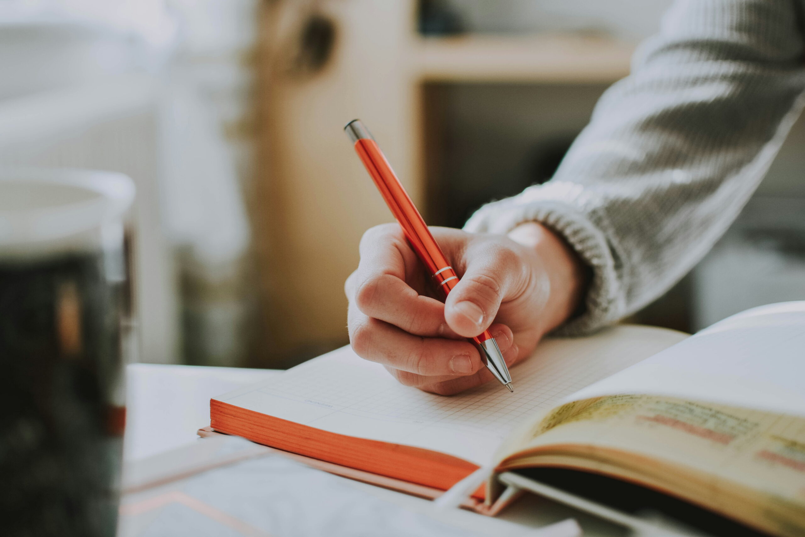 A close-up view of a person wearing a grey sweater holding a vibrant orange pen, actively writing on the grid-lined pages of an open notebook.