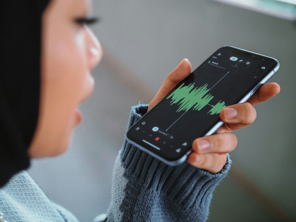 Close-up profile of a person holding a smartphone close to their mouth, recording voice audio displayed as a green soundwave on screen.