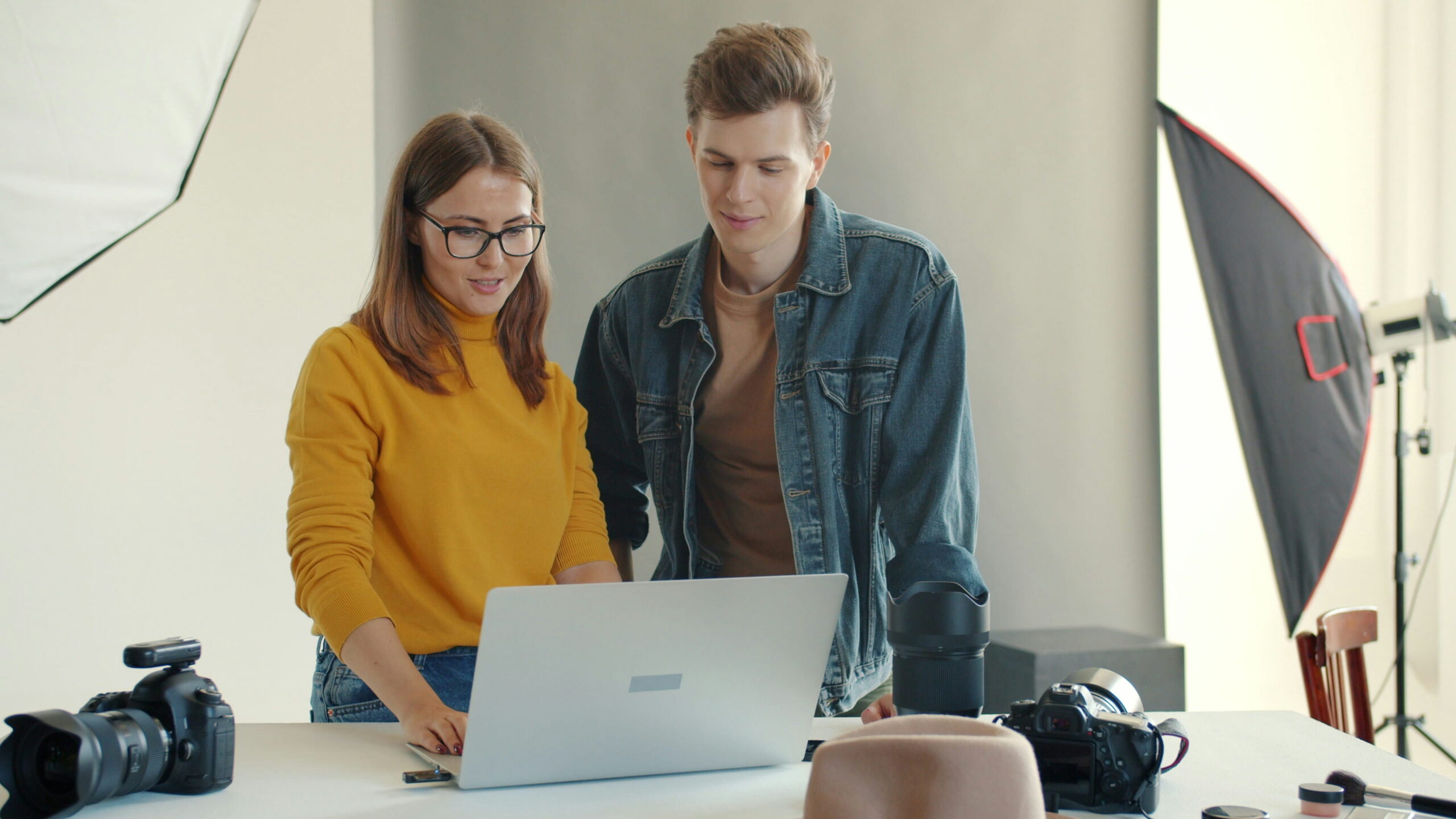 A man and a woman standing in a photography studio, smiling as they look at photos on an open laptop placed on a white table surrounded by cameras, lenses, and lighting equipment.