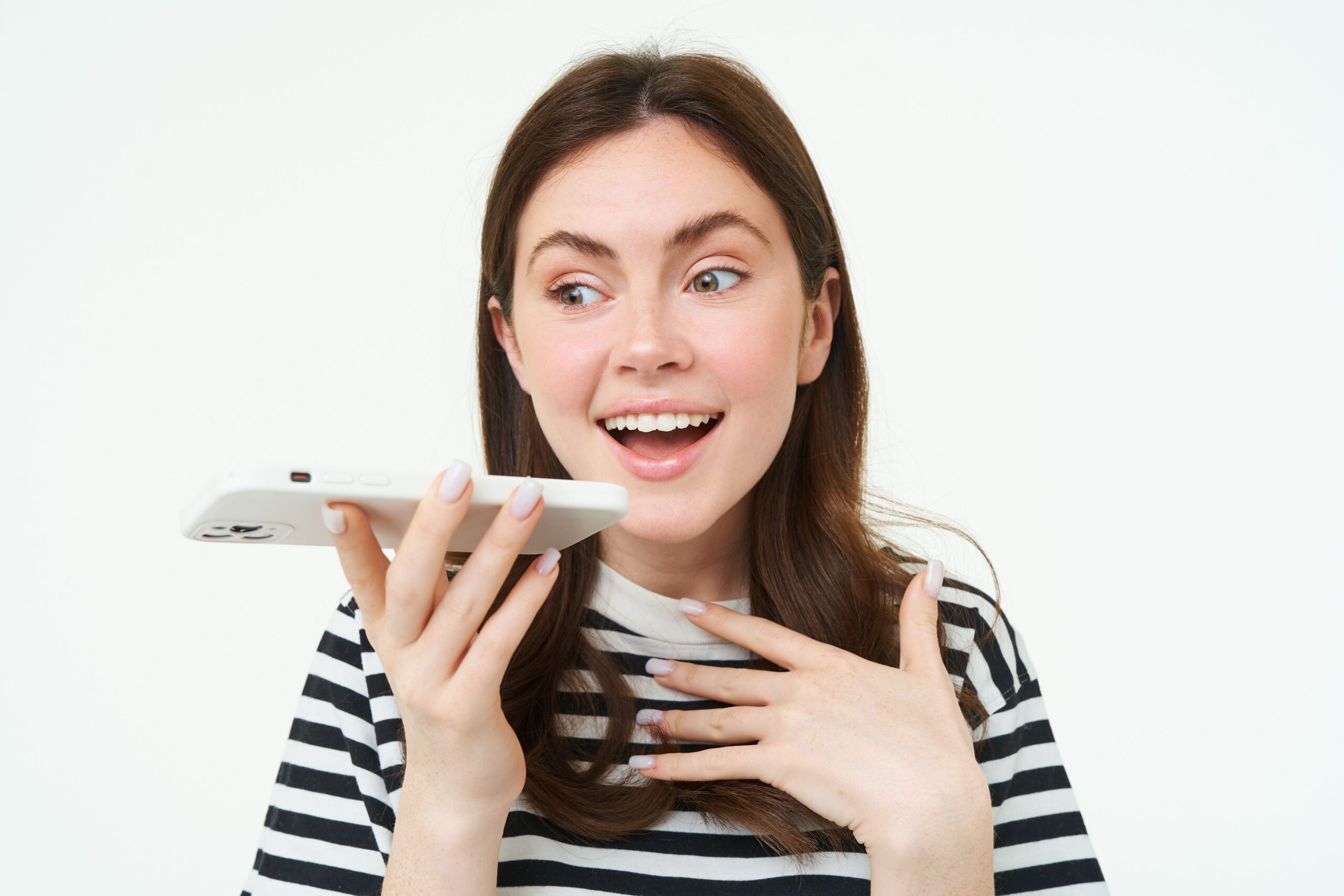 A smiling brunette woman in a striped shirt holding a mobile phone near her mouth to record audio or use speakerphone against a white background.