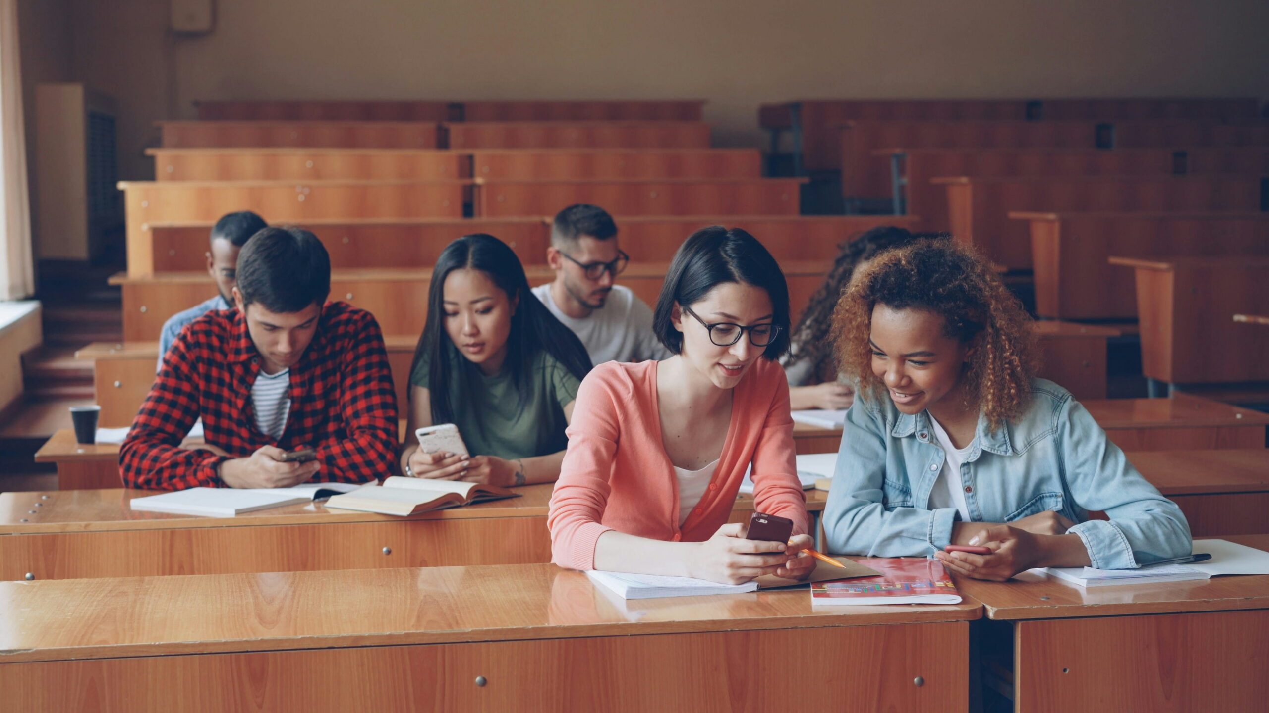 A group of diverse university students sitting at wooden desks in a large lecture hall, looking down at and using their smartphones.