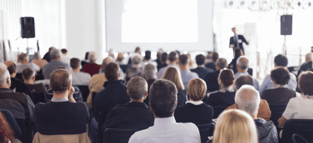 A seated audience facing a speaker and projection screen in a conference hall.