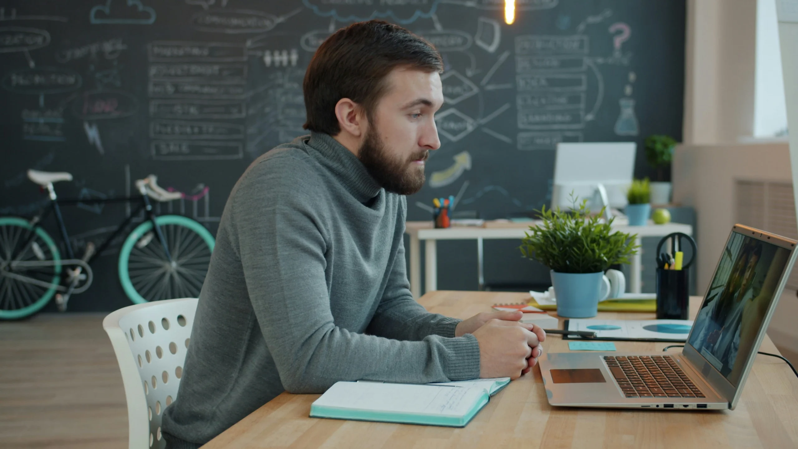 A bearded man in a gray turtleneck sitting at a desk with a laptop and a notebook, attentively participating in a virtual online class in a modern workspace.
