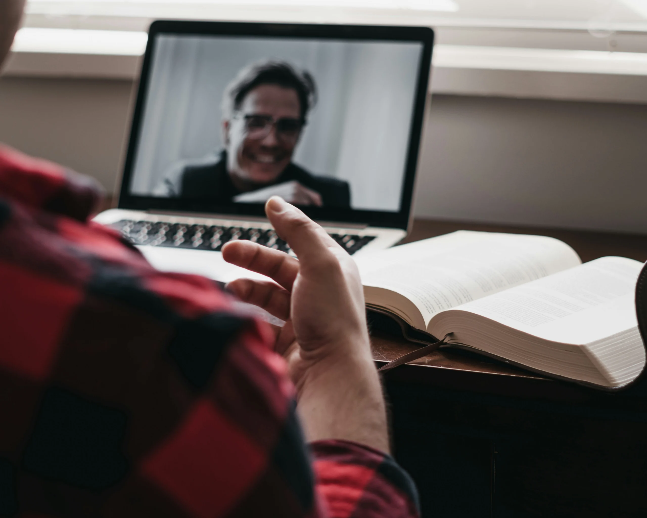 Over-the-shoulder view of a student in a red plaid shirt studying at a desk with an open textbook while attending a live online class or video call on a laptop.