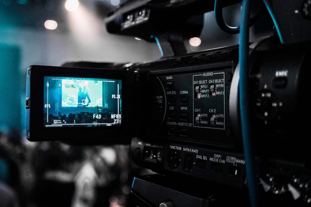 Professional broadcast video camera featuring a flip-out monitor displaying live event footage and comprehensive side-panel controls for audio routing, gain adjustment, and camera functions.