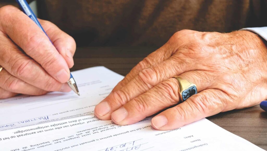 An older person's hands filling out a Dutch document with a blue pen, wearing a signet ring.