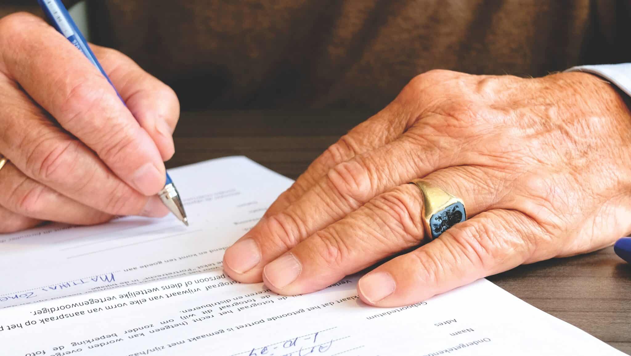 An older person's hands filling out a Dutch document with a blue pen, wearing a signet ring.