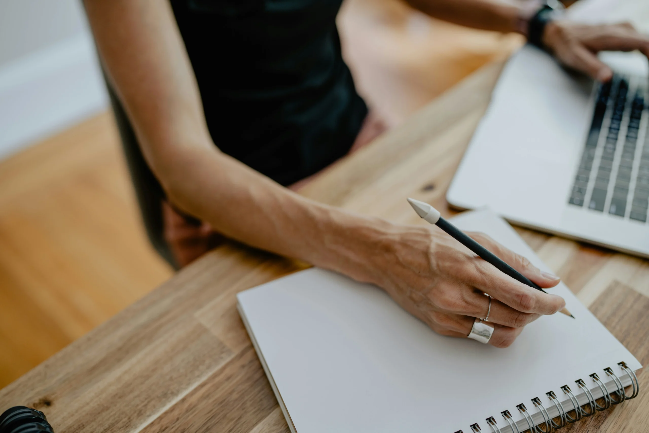 Close-up of a person writing notes in a spiral notebook next to a laptop on a wooden desk.