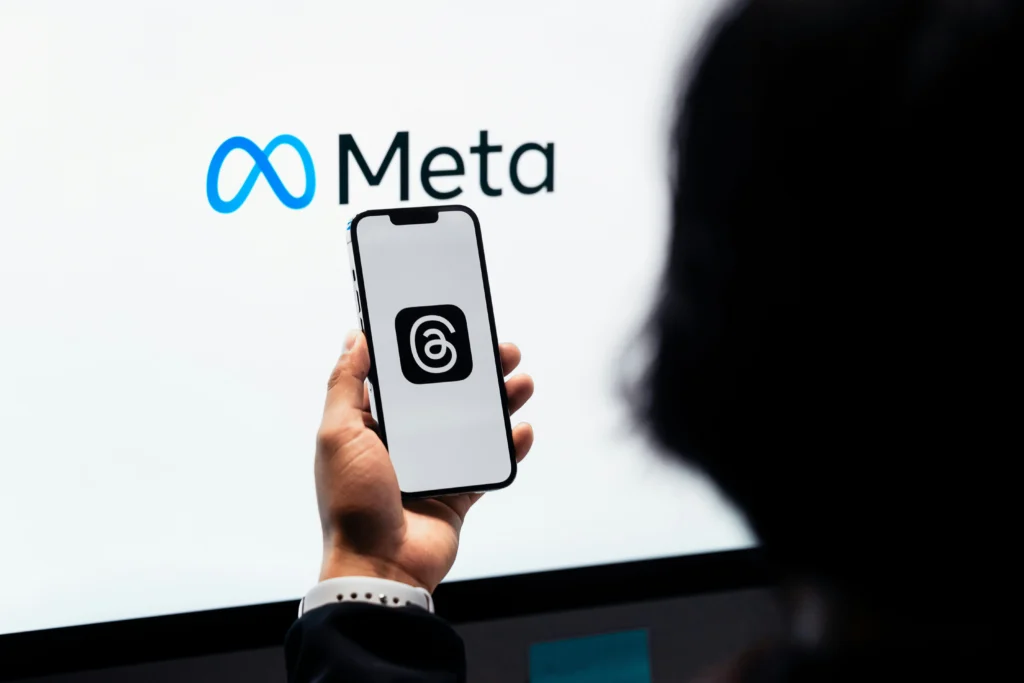 A person's hands holds a mobile device with a screen behind the phone showing the Meta logo on it.