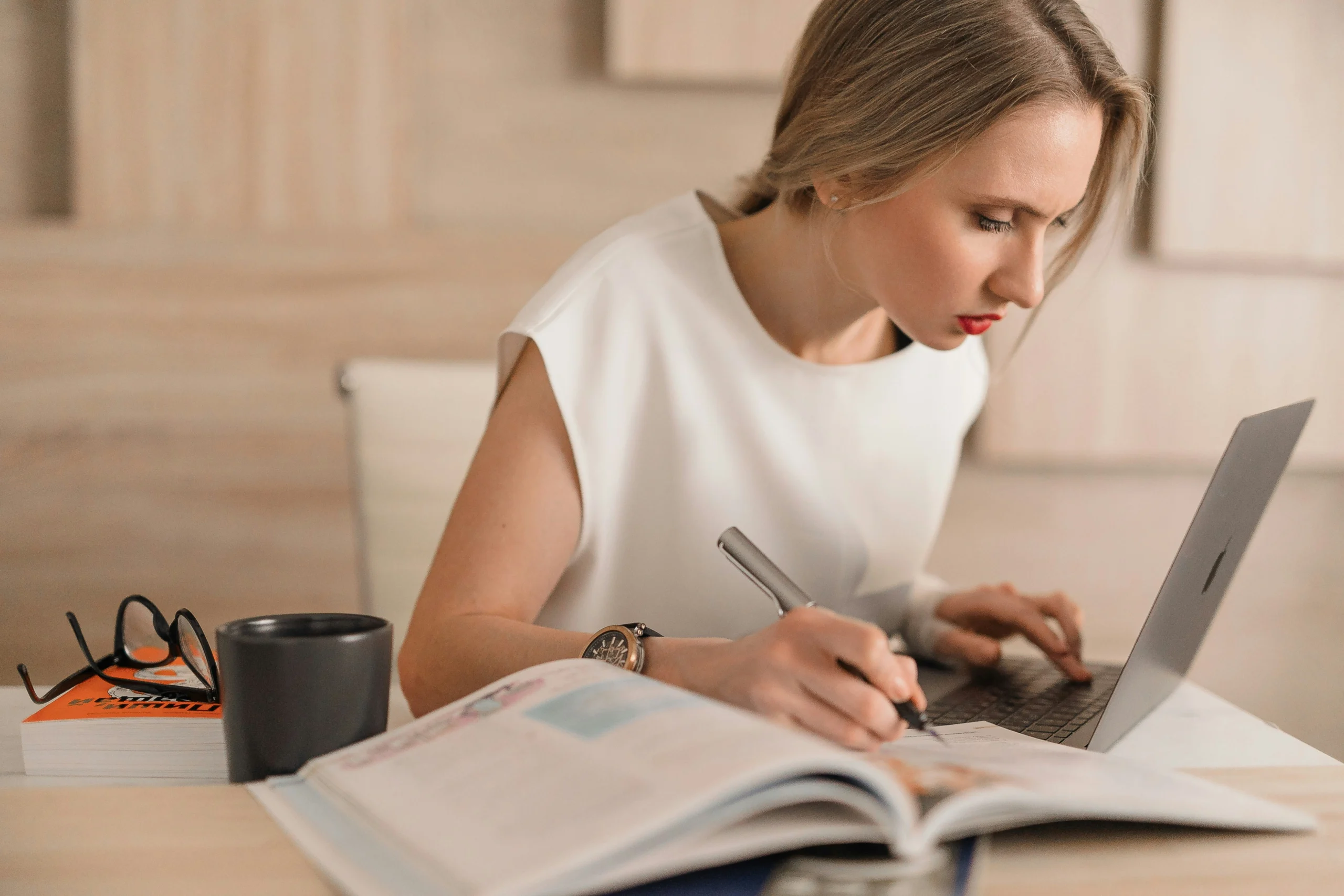 A young blonde woman wearing a white sleeveless top sits at a wooden desk, deeply concentrated as she writes in an open textbook with a silver pen. Her right hand rests on the keyboard of a partially visible silver laptop. In the foreground, there are eyeglasses and a black mug. The background consists of light beige wooden panels.