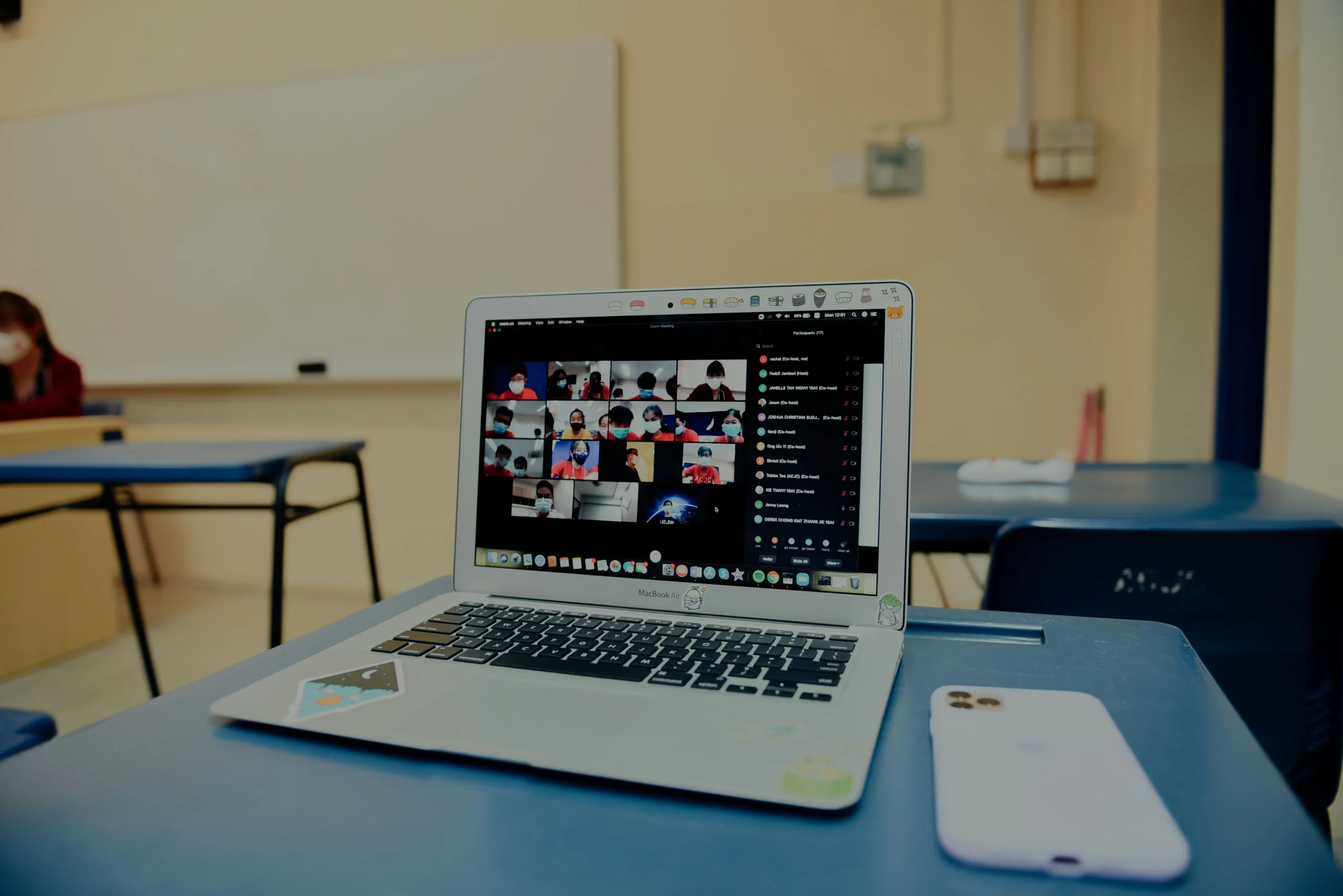 A silver laptop sits on a blue desk in an empty classroom. The screen displays a Zoom call with multiple participants in a grid view. Next to the laptop is a white smartphone. In the background, there is a whiteboard and another desk.