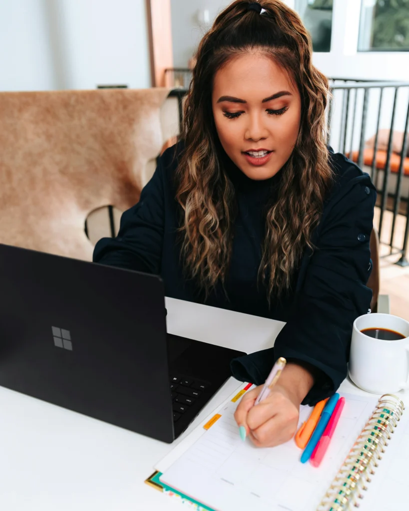 A female student with wavy hair and a black shirt learns from a Microsoft Surface Laptop 3 in Black and is shown writing in colored markers as she studies.