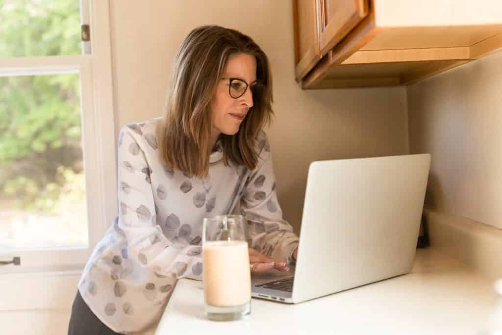 woman standing beside a table with a glass of milk looking at her laptop