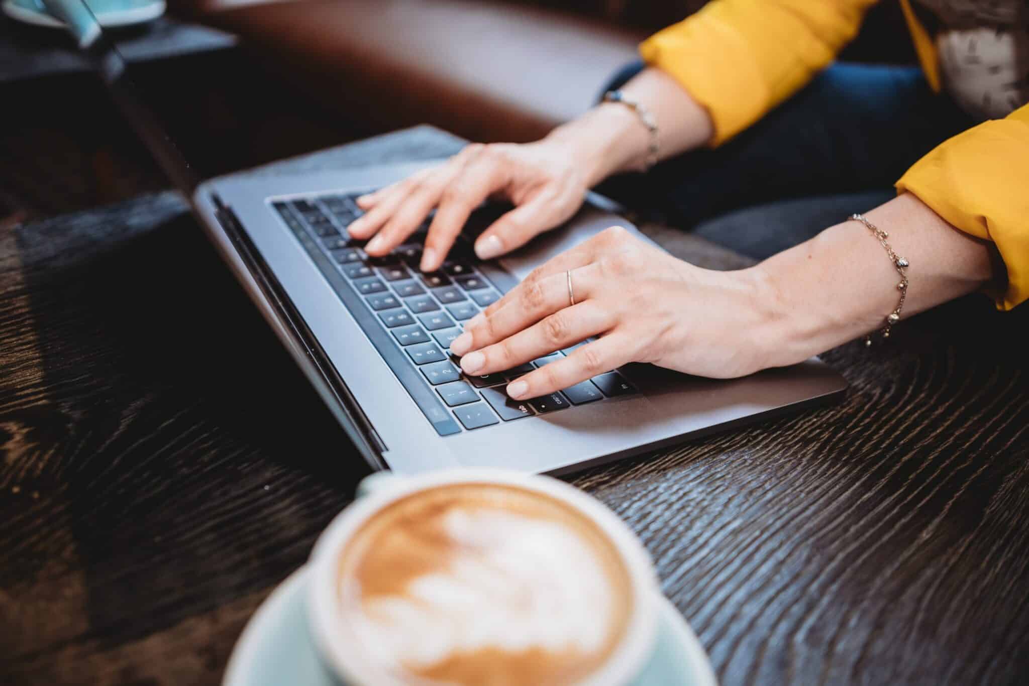 a persons hands typing on a laptop with a cup of coffee beside the laptop