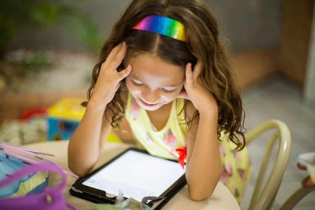 a young girl wearing a rainbow headband working on her tablet on a table