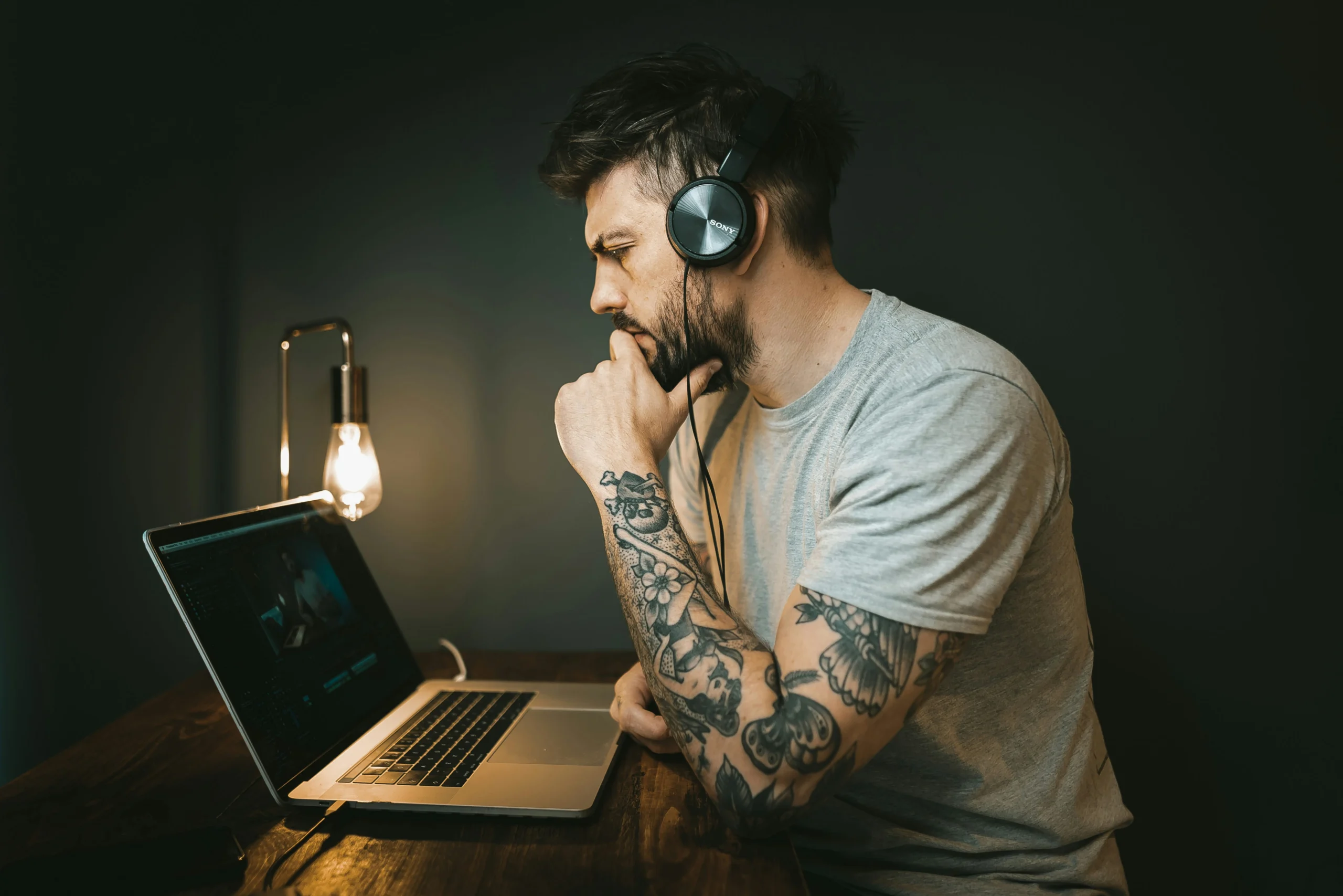 A tattooed man wearing headphones and a gray t-shirt sitting at a wooden desk in a dimly lit room, intently focused on his open laptop with a small desk lamp glowing in the background.