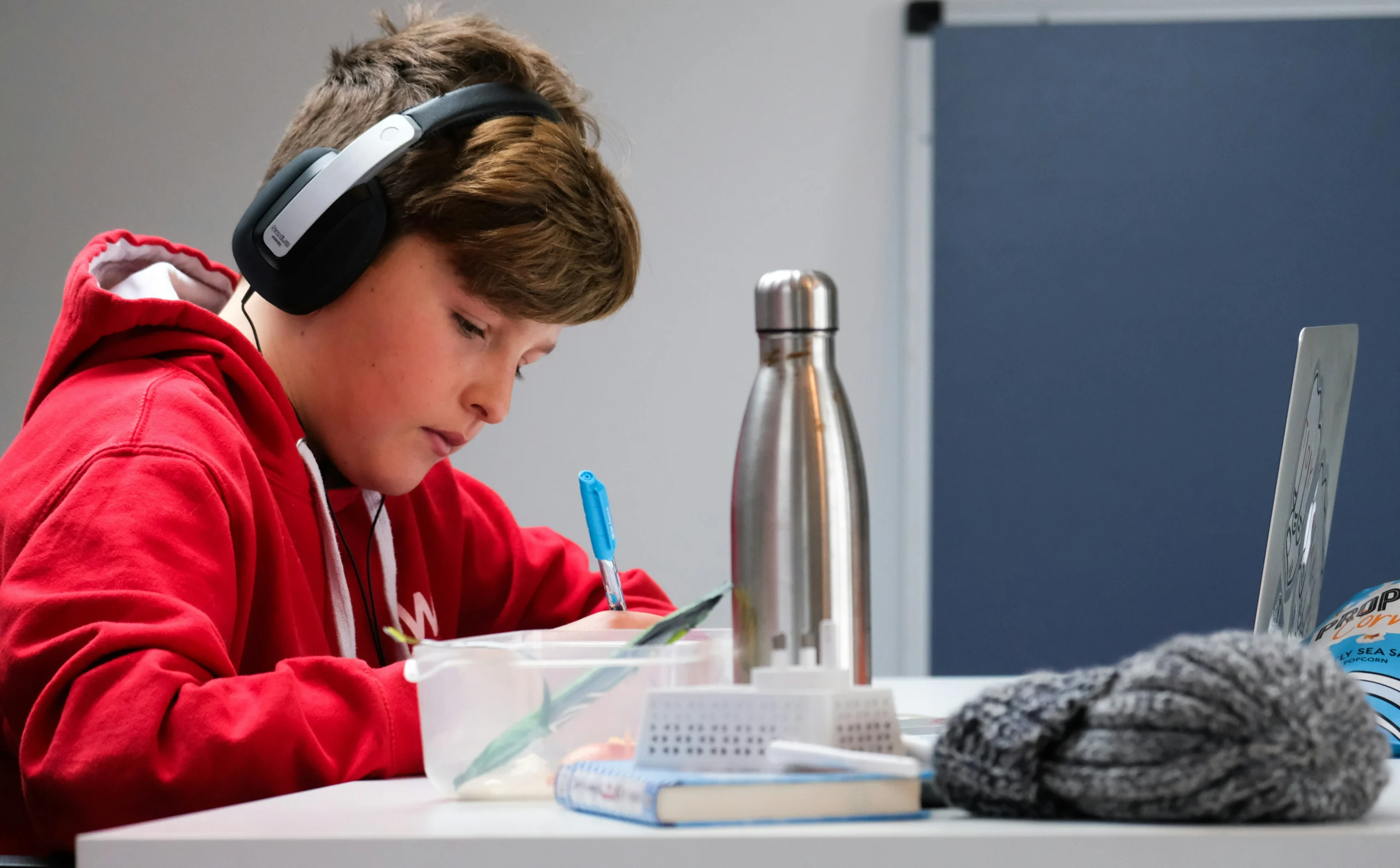 A young boy wearing a red hoodie and black over-ear headphones, intently focused on writing or drawing in a container at a white desk with a metal water bottle, a laptop, and various desk items nearby.