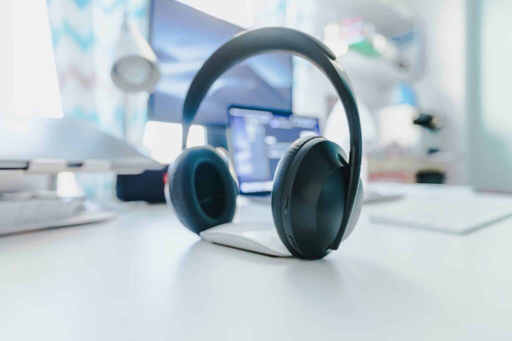 A pair of black over-ear wireless headphones resting upright on a white desk, with a blurred home office setup featuring a computer monitor and lamp in the background.