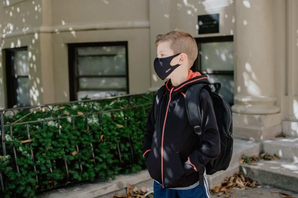 child wearing aa facemask with his backpack on his back standing in front of a building