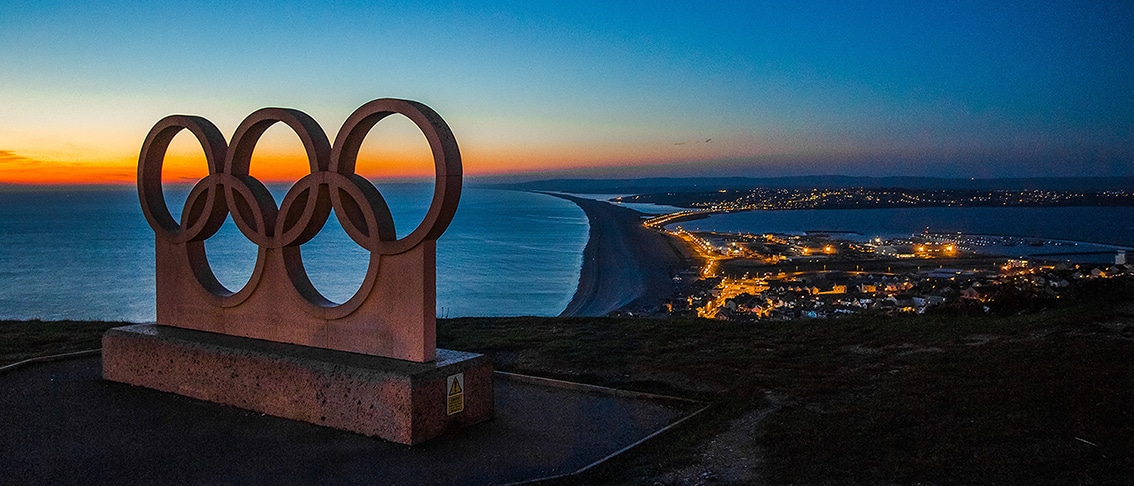Image of a stone statue of the Olympic interlaced rings symbol, with the ocean near a city in the background.