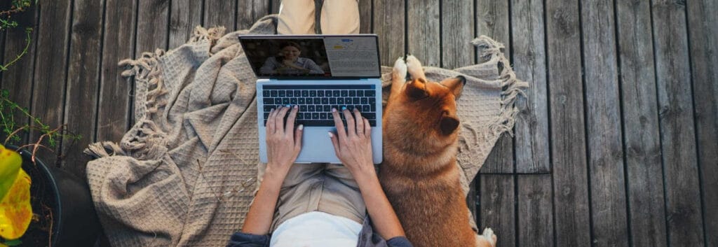 person typing webex transcription on his laptop with a dog beside him on a porch