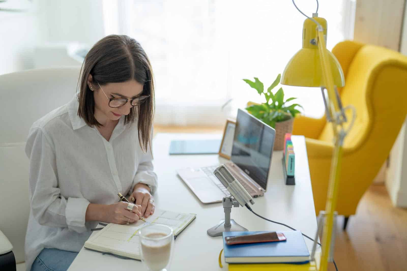 woman writing notes on a notebook on her desk with her laptop and mic