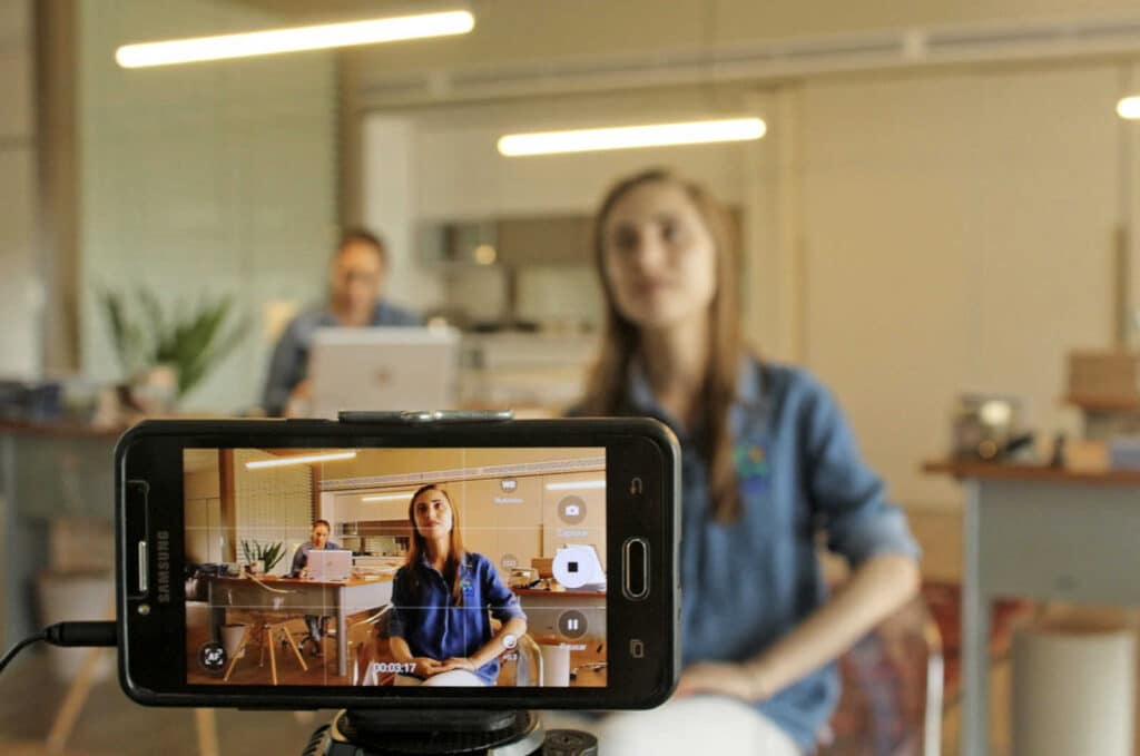 a mobile phone capturing the video of a woman in the background sitting on a chair