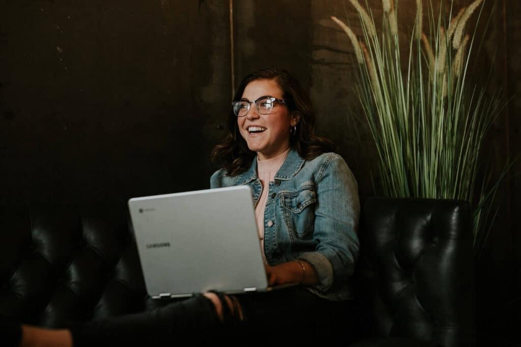 smiling woman wearing her glasses with her laptop on her lap and a pot of plant at her back
