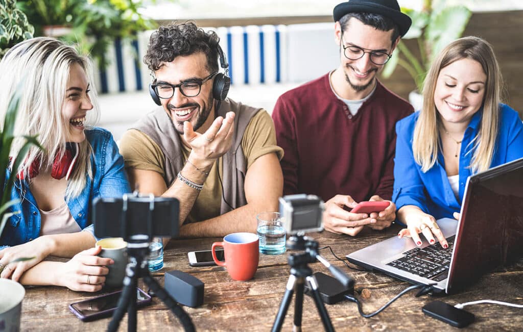 Four people - two men and two women - sit at a table. Open laptops, cameras and video equipment are on the table in front of them.