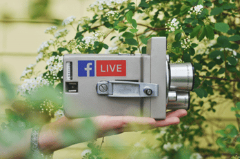 a camera with facebook live logo being held by a hand with leaves on the background