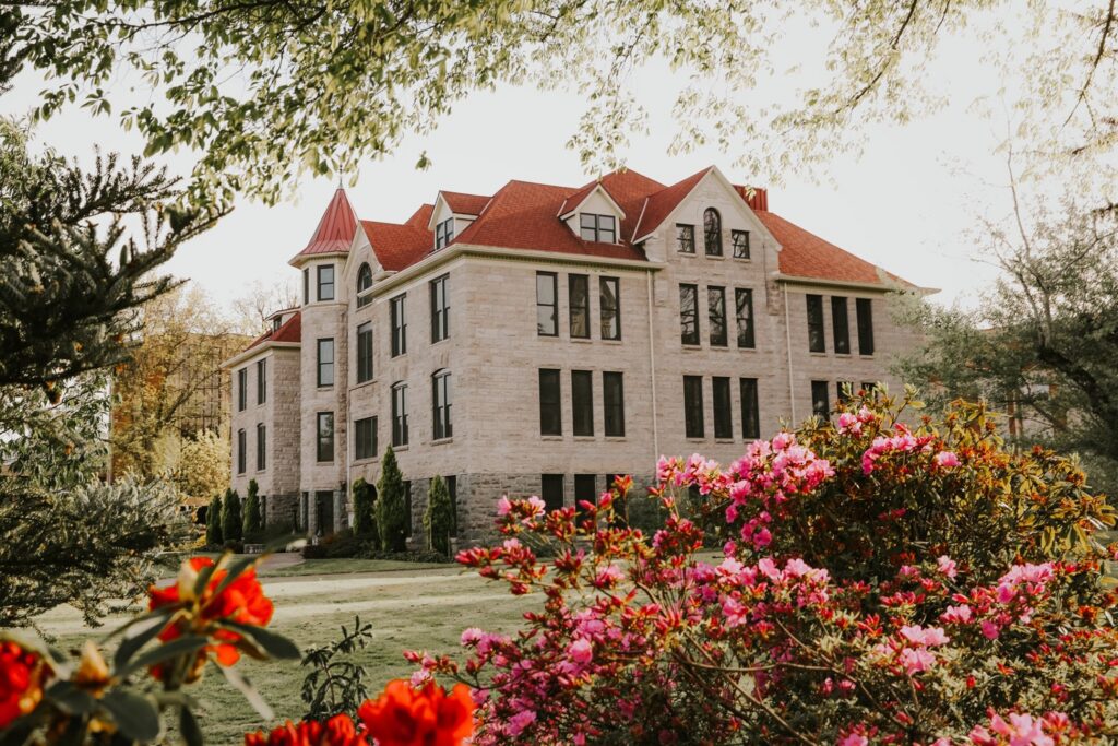 Elegant stone building with red gabled roofs surrounded by blooming pink and red flowers and lush greenery, photographed in warm morning light.