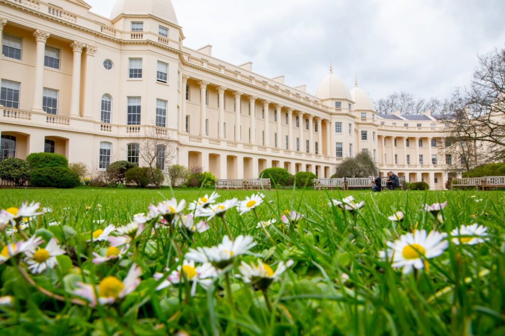 Elegant neoclassical building with cream-colored walls, tall columns, and domed towers, viewed from a lawn dotted with white daisies under a cloudy sky.