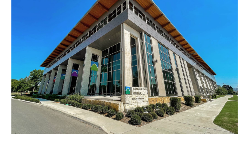 Modern Alamo Colleges District building with tall glass windows, concrete columns, and a wooden roof overhang, surrounded by landscaped greenery under a clear blue sky.