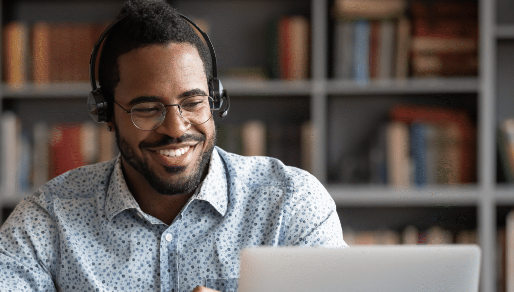 Smiling man wearing glasses and a headset working on a laptop in front of a bookshelf filled with books, suggesting remote learning or virtual collaboration.