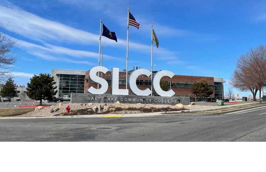Entrance sign of Salt Lake Community College featuring large white SLCC letters and three flagpoles flying the U.S., Utah, and college flags in front of a modern brick and glass building under a bright blue sky.