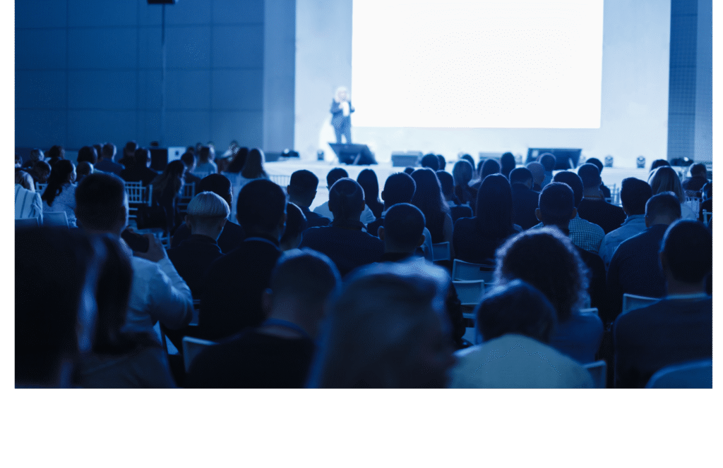 Audience seated in a large conference hall watching a speaker on stage in front of a bright screen, illuminated by blue lighting.