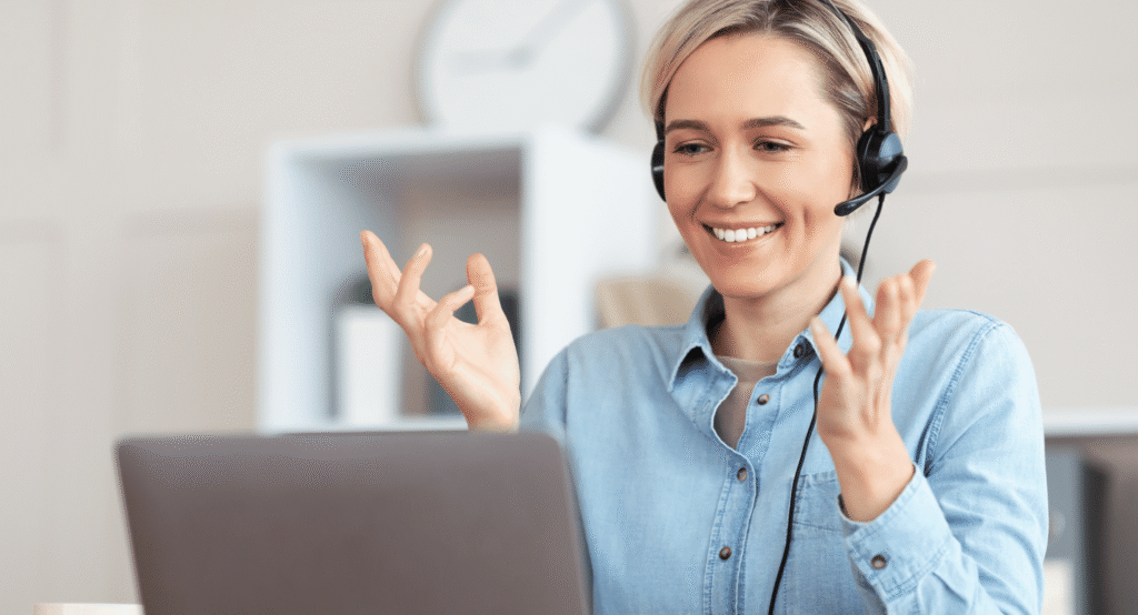 Smiling woman wearing a headset engaged in a video call, gesturing with her hands while sitting at a desk with a laptop in a bright home office.