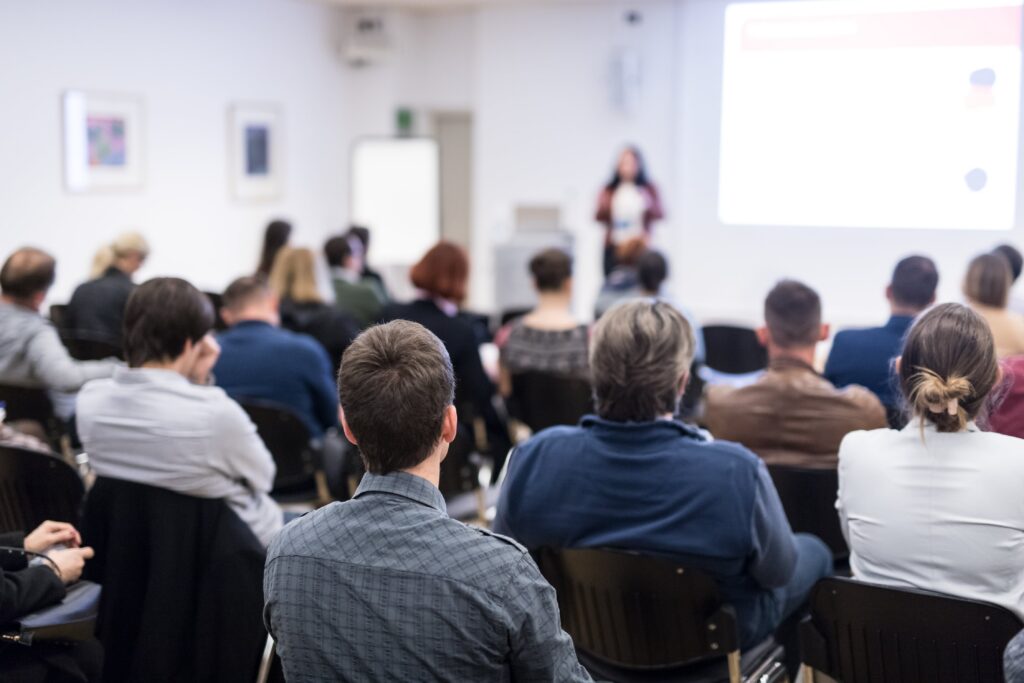 An in-person lecture taking place with the picture taken from the back of the room showing the backs of attendees