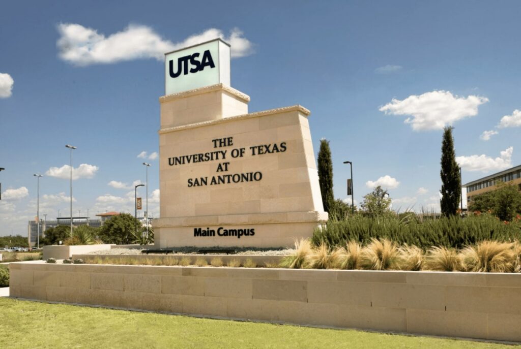 Stone monument sign for The University of Texas at San Antonio main campus, featuring bold UTSA lettering under a clear blue sky with landscaped greenery and tall trees.