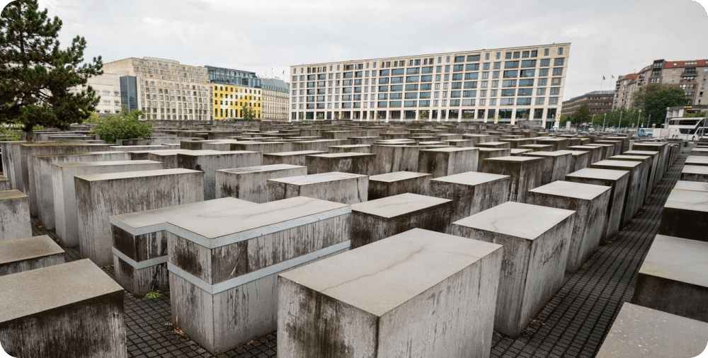Image showcasing a building in the background with memorial of Holocaust in the foreground of large square grey cement stones.