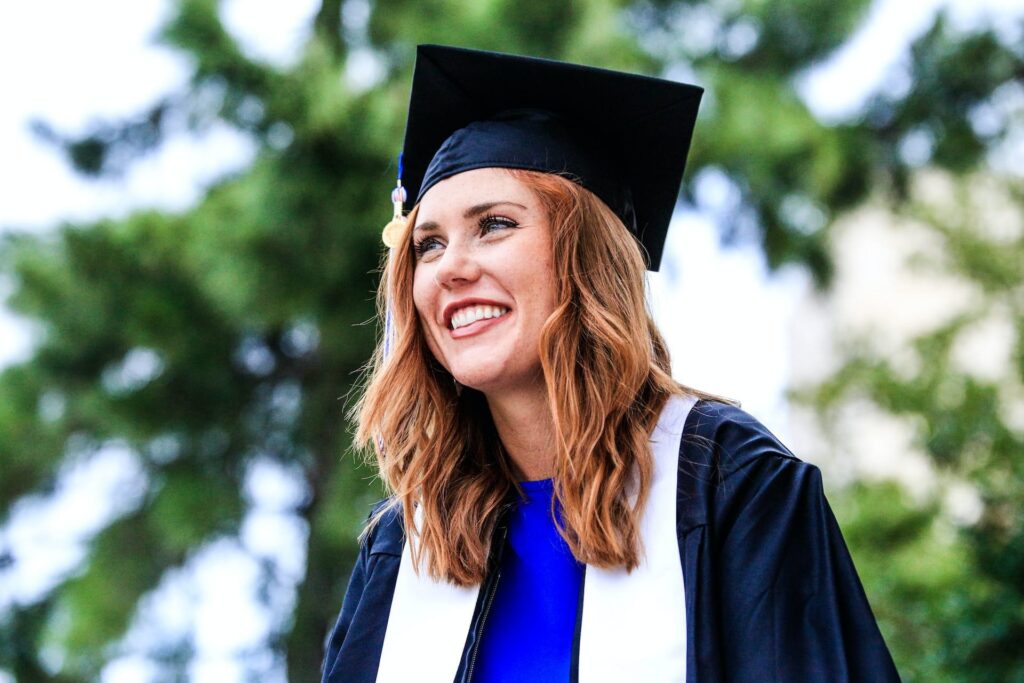 Smiling graduate wearing a black cap and gown with a white stole and blue dress, standing outdoors with green trees in the background.