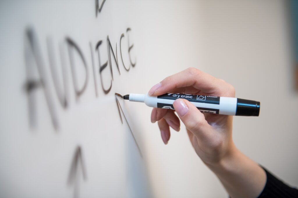 Person's hand shown drawing on white board holding black dry erase pen with the word 'Audience' shown drawn on the board.