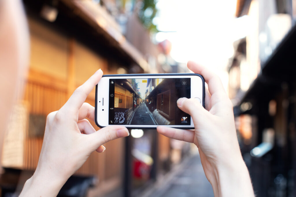 Person holding a smartphone horizontally to photograph a narrow alley lined with traditional wooden buildings; the phone screen shows the same scene in focus while the background appears blurred.