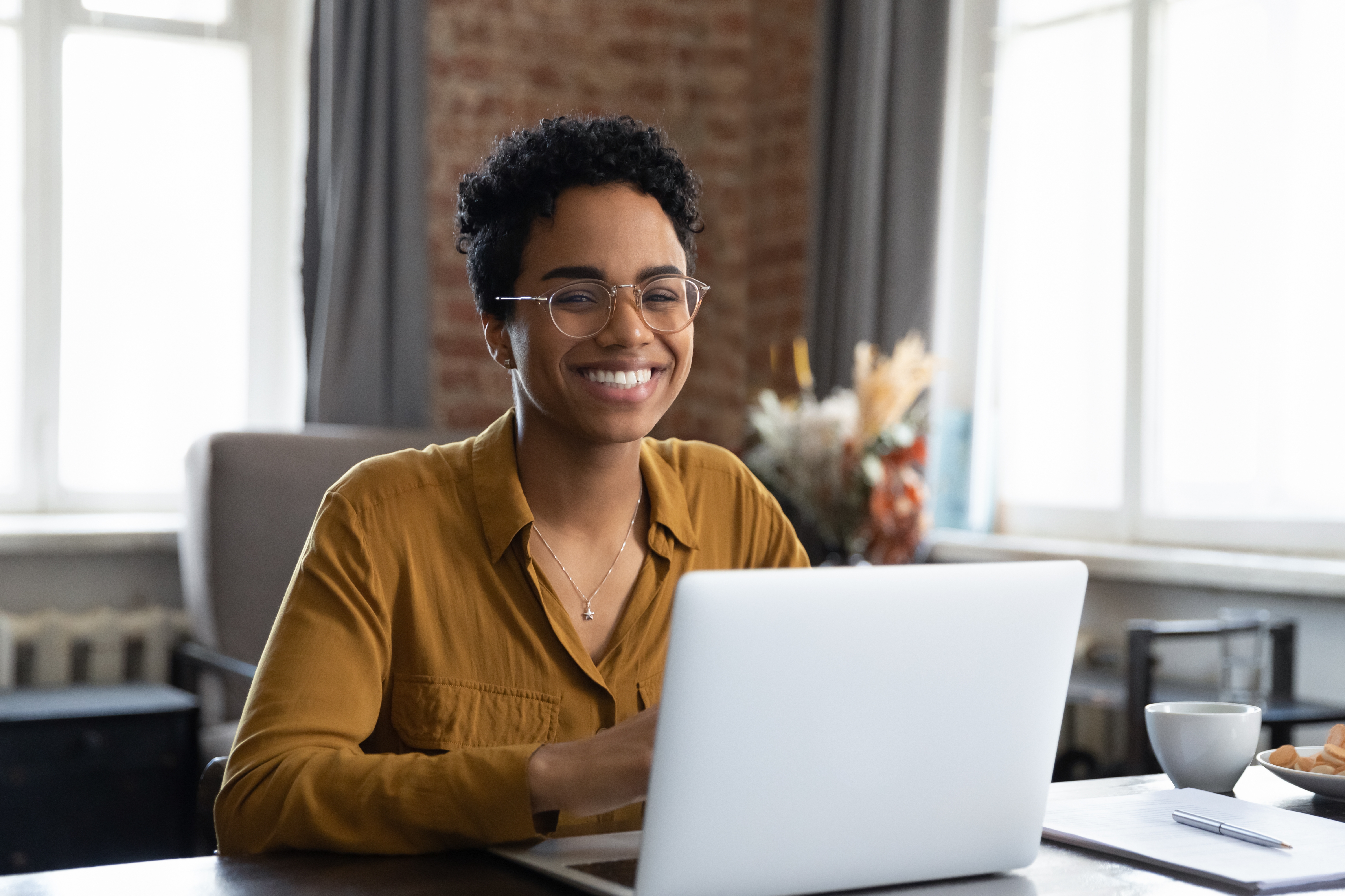 woman wearing glasses and smiling sits at a desk working on a computer
