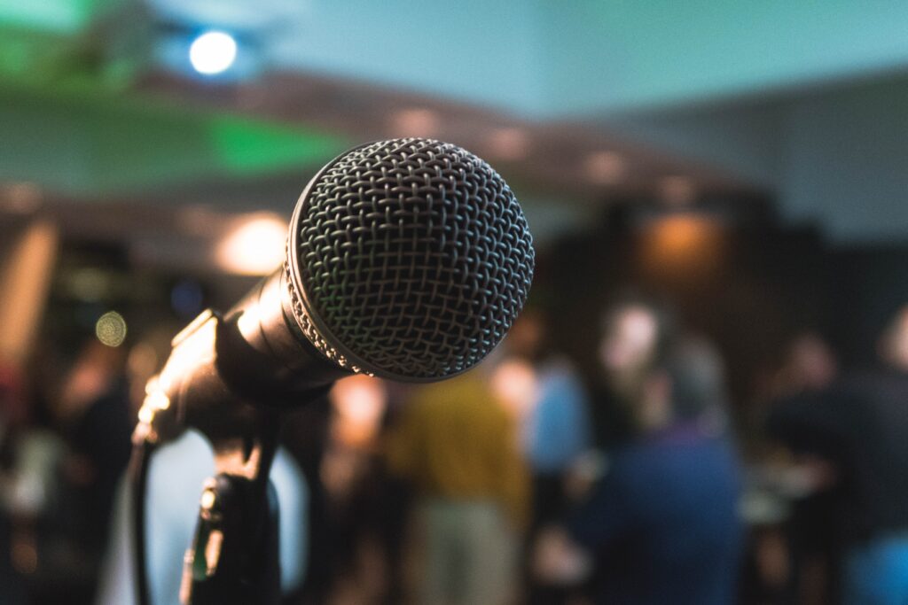A microphone appears close up on a stage with a blurred image of people attending a conference to watch from their seats