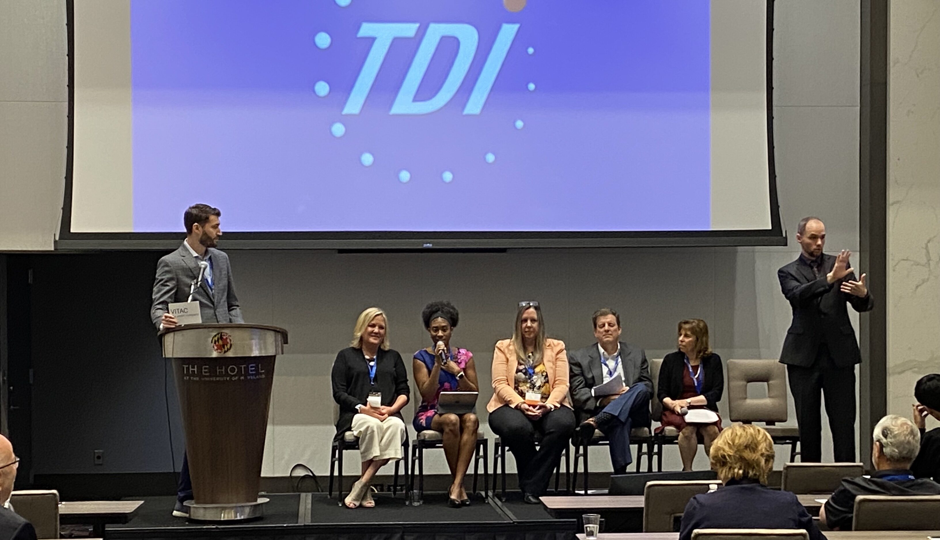 Matthew Schwartz of Verbit moderates an event at TDI, Heather York, Crystal Evans, Stacey Romero, Larry Walke and Darlene Parker are sitting and participating while an ASL interpreter signs to the audience