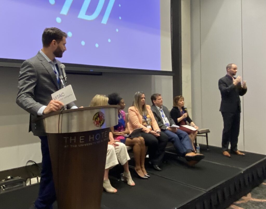 Matthew Schwartz of Verbit moderates an event at TDI, Heather York, Crystal Evans, Stacey Romero, Larry Walke and Darlene Parker are sitting and participating while an ASL interpreter signs to the audience 