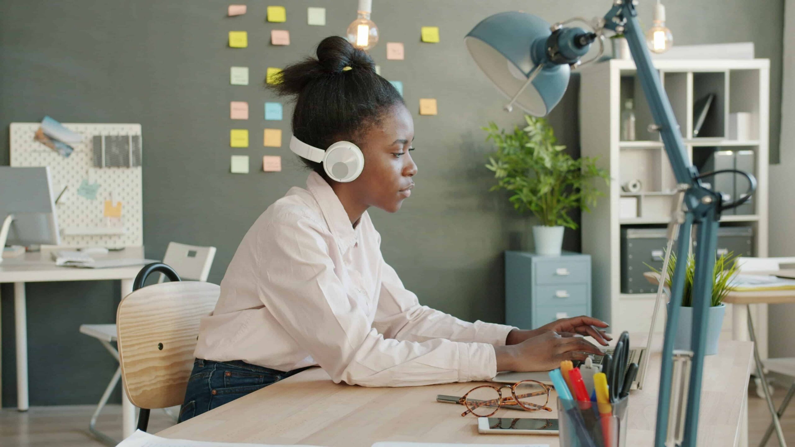 A woman in a white blouse sits working from her desk on a laptop with headphones around her ears