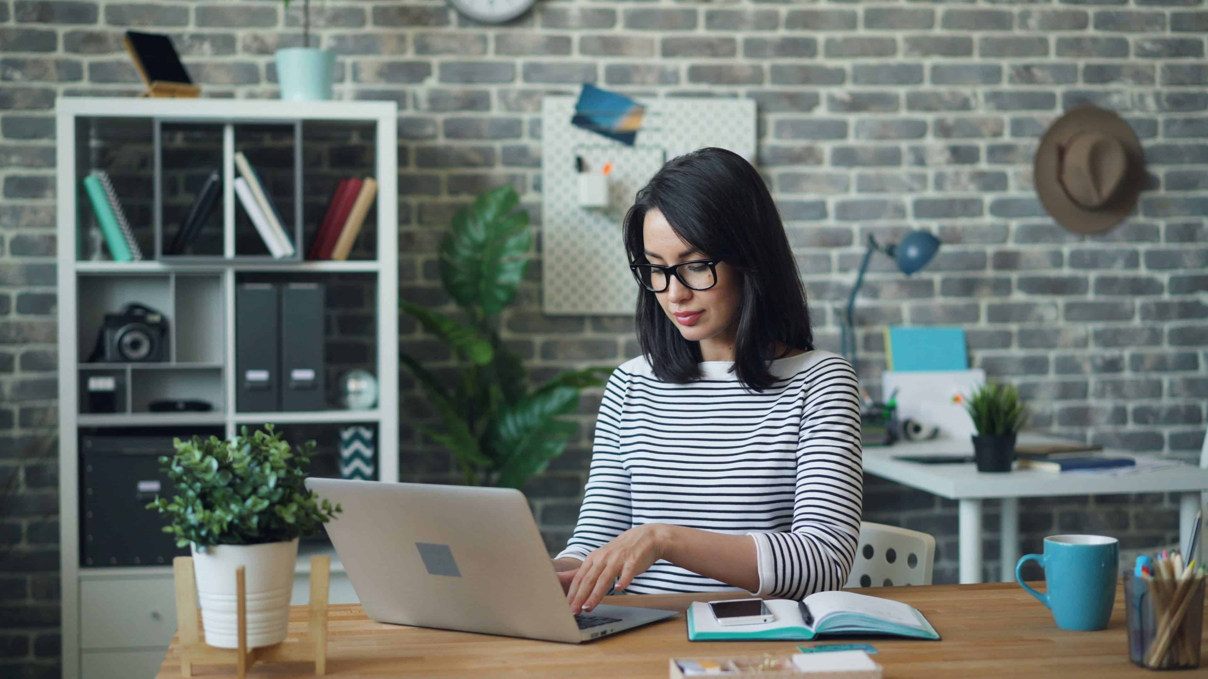 A woman with short brown hair and glasses sits working from her laptop with brick wall and shelves visible behind her
