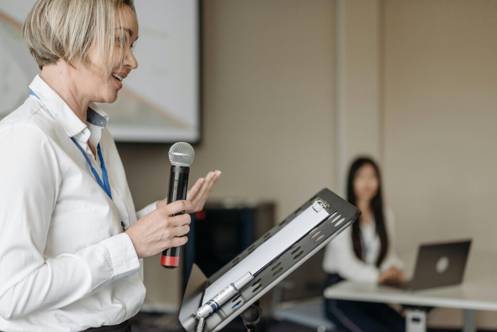 Woman standing at podium, speaking to classroom.